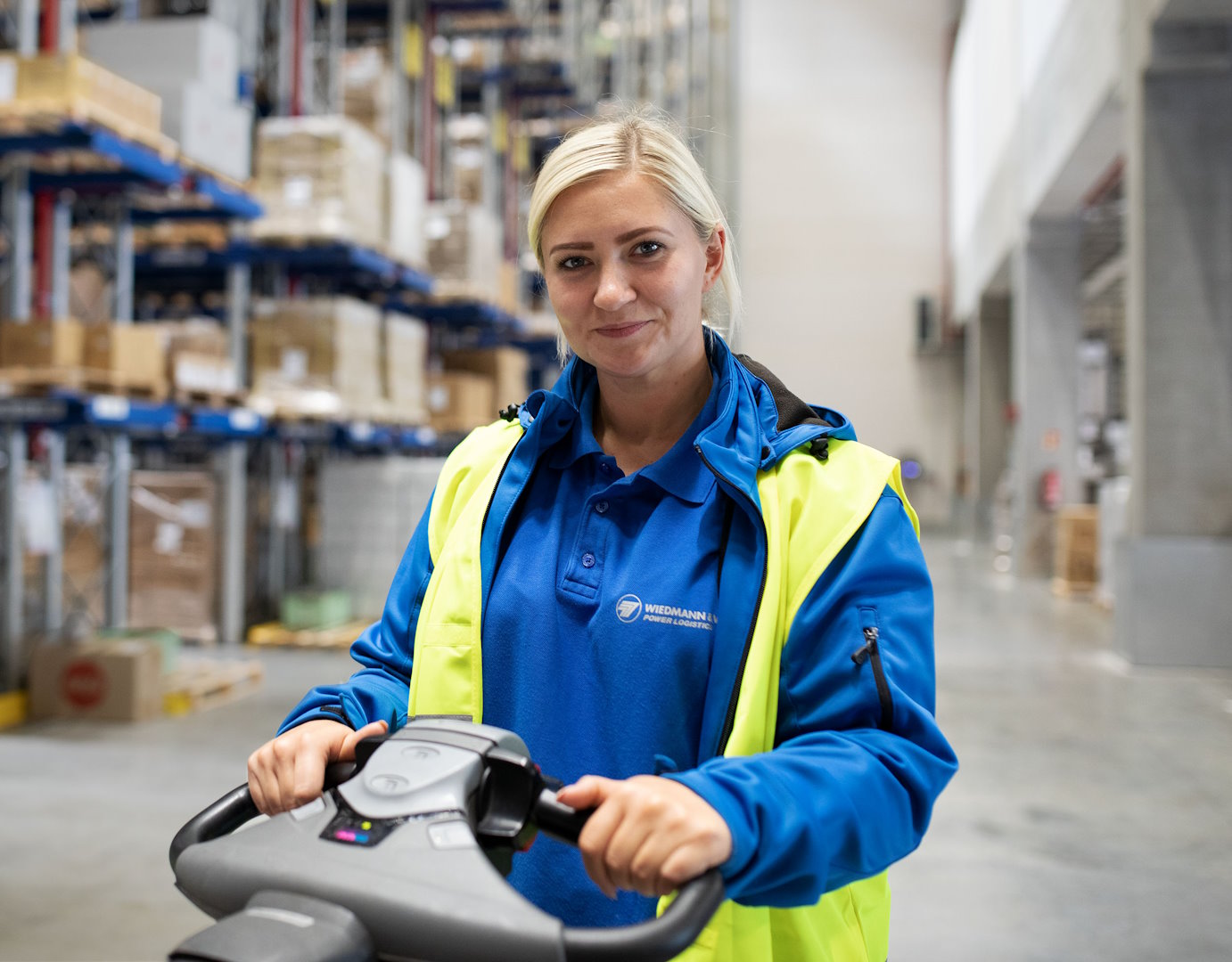 Ein Frau mit blauer Jacke und gelber Warnweste steht auf einer Elektroameise in einem Logistikzentrum.