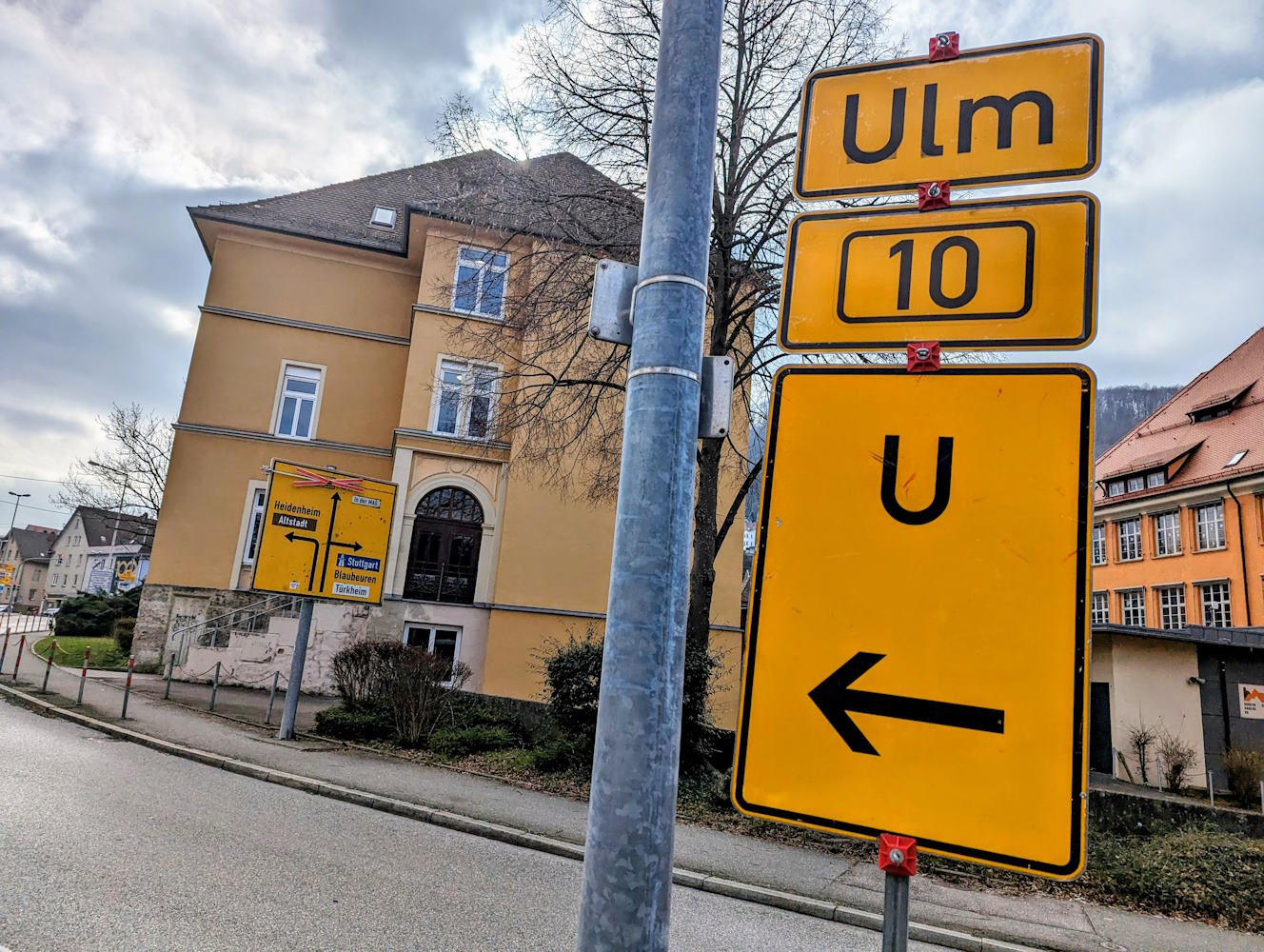 Drei gelbe Schilder übereinander stehen vor einer Straße.