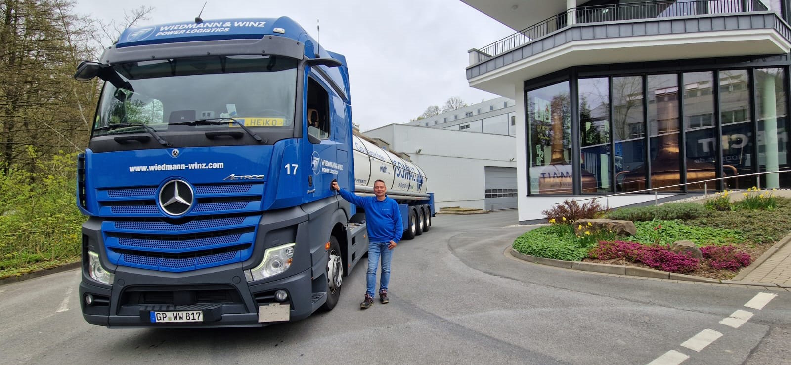 Ein Fahrer steht neben seinem blauen LKW mit einem weißen Tankauflieger vor einem modernen Gebäude.